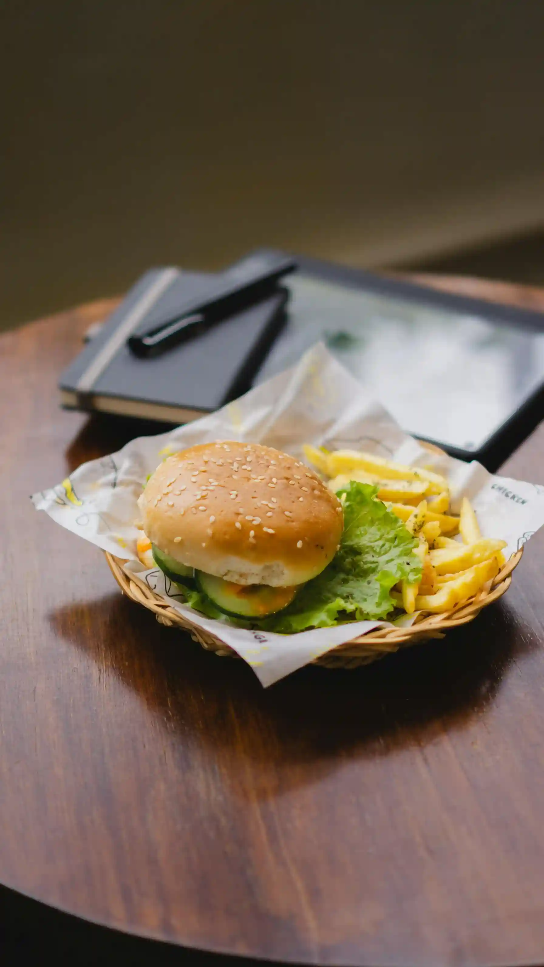 A sesame seed burger and fries served in a woven basket lined with a custom food basket liner, placed on a wooden table alongside a menu and a pen