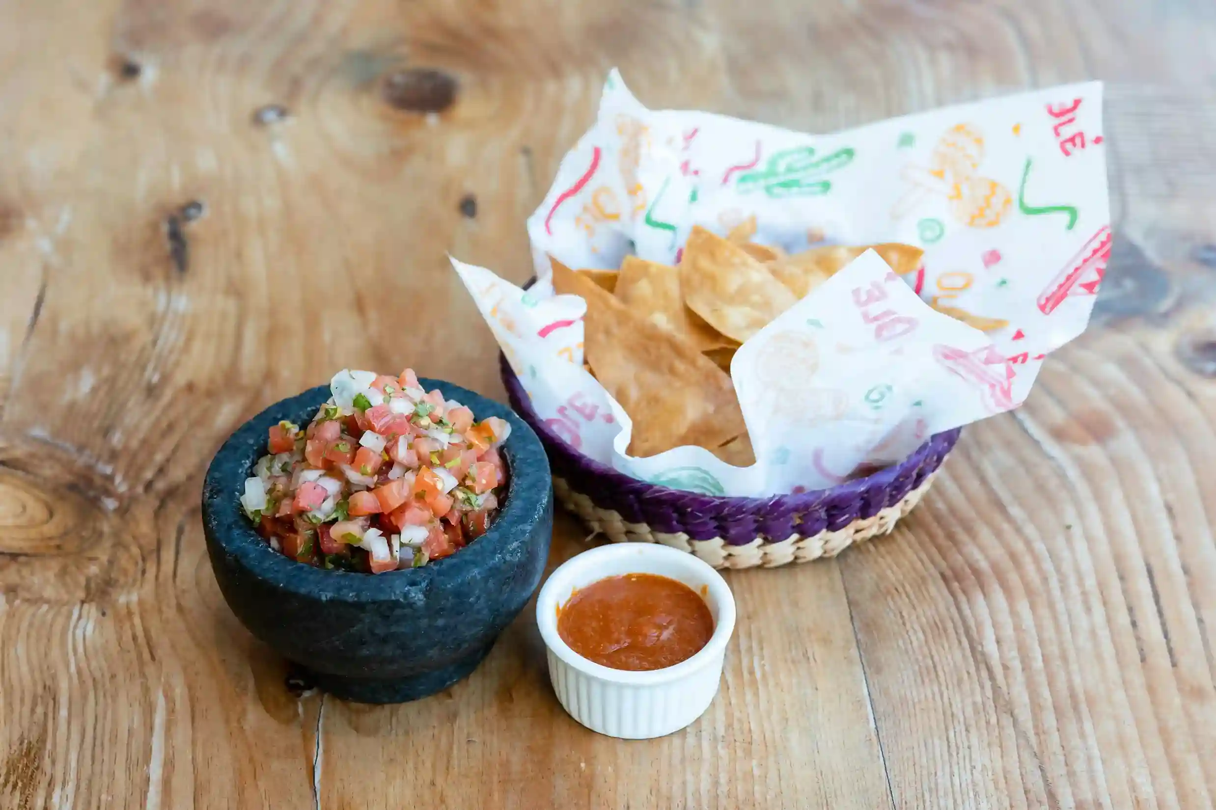 Crispy tortilla chips served in a woven basket lined with a custom-printed food basket liner, accompanied by fresh salsa in a stone bowl and spicy dip in a ramekin, placed on a wooden table