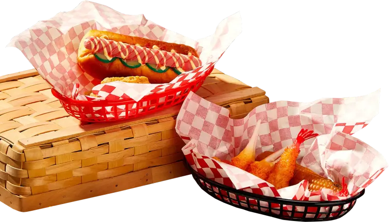 Two red food baskets lined with custom-printed red-and-white checkered basket liners. One basket holds a hot dog topped with mustard, ketchup, and sliced cucumbers, while the other contains fried shrimp and a mini corn cob, placed on a woven wooden box