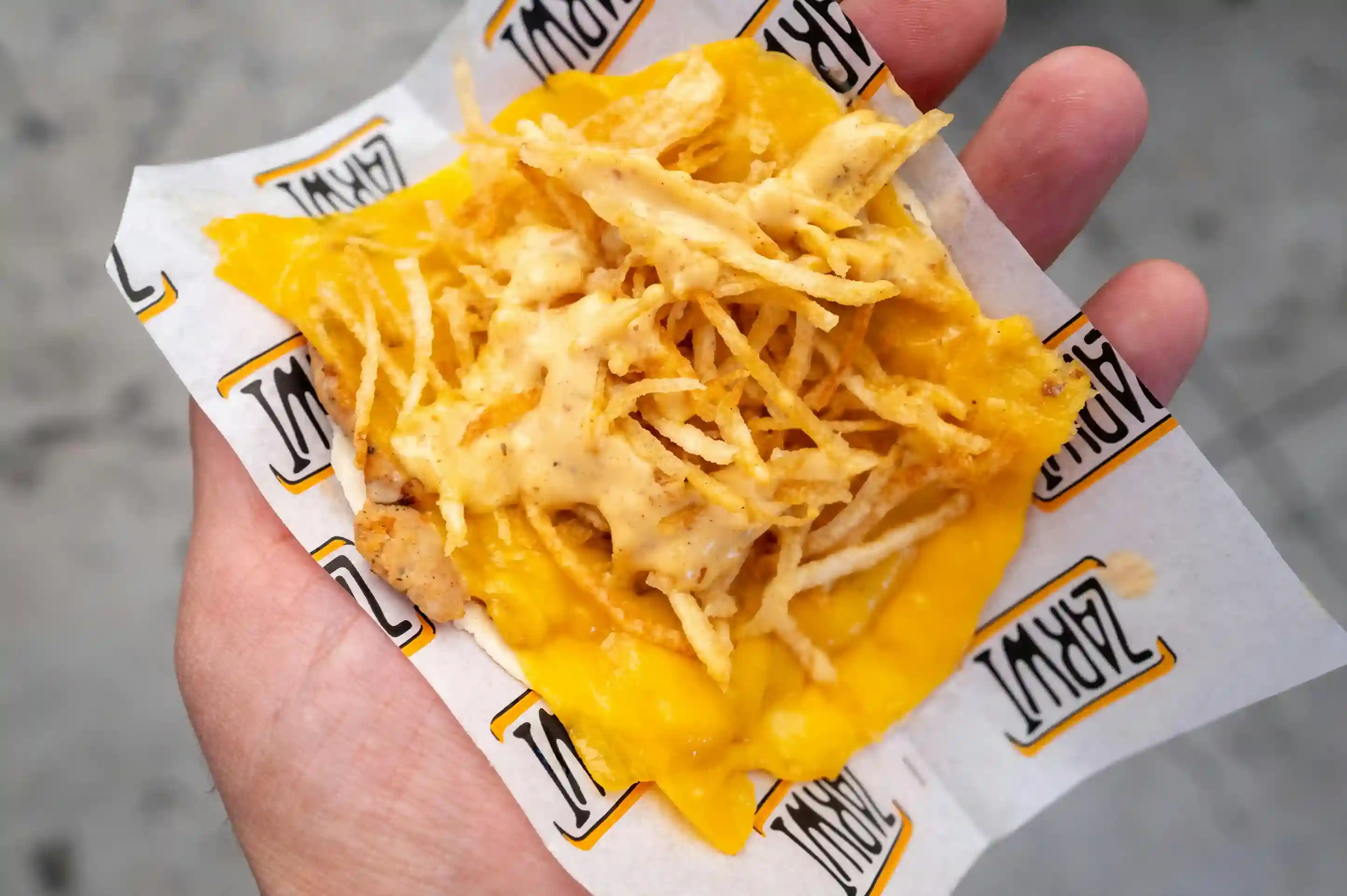 Hand holding cheesy nachos with crispy toppings served on a custom-printed cheese paper featuring a branded design