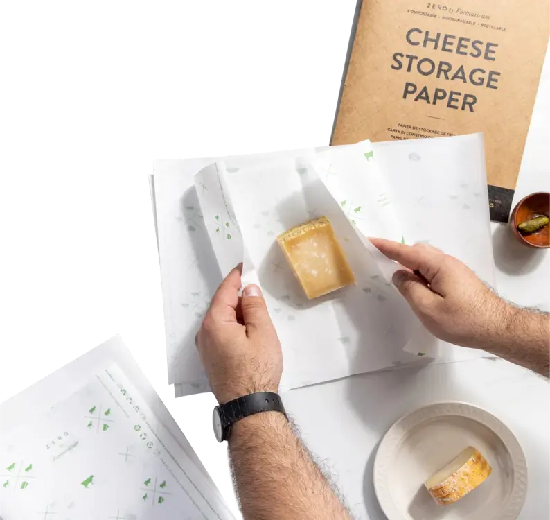 Hands wrapping a block of cheese in custom cheese storage paper, with a cheese storage paper package and a small plate of cheese in the background