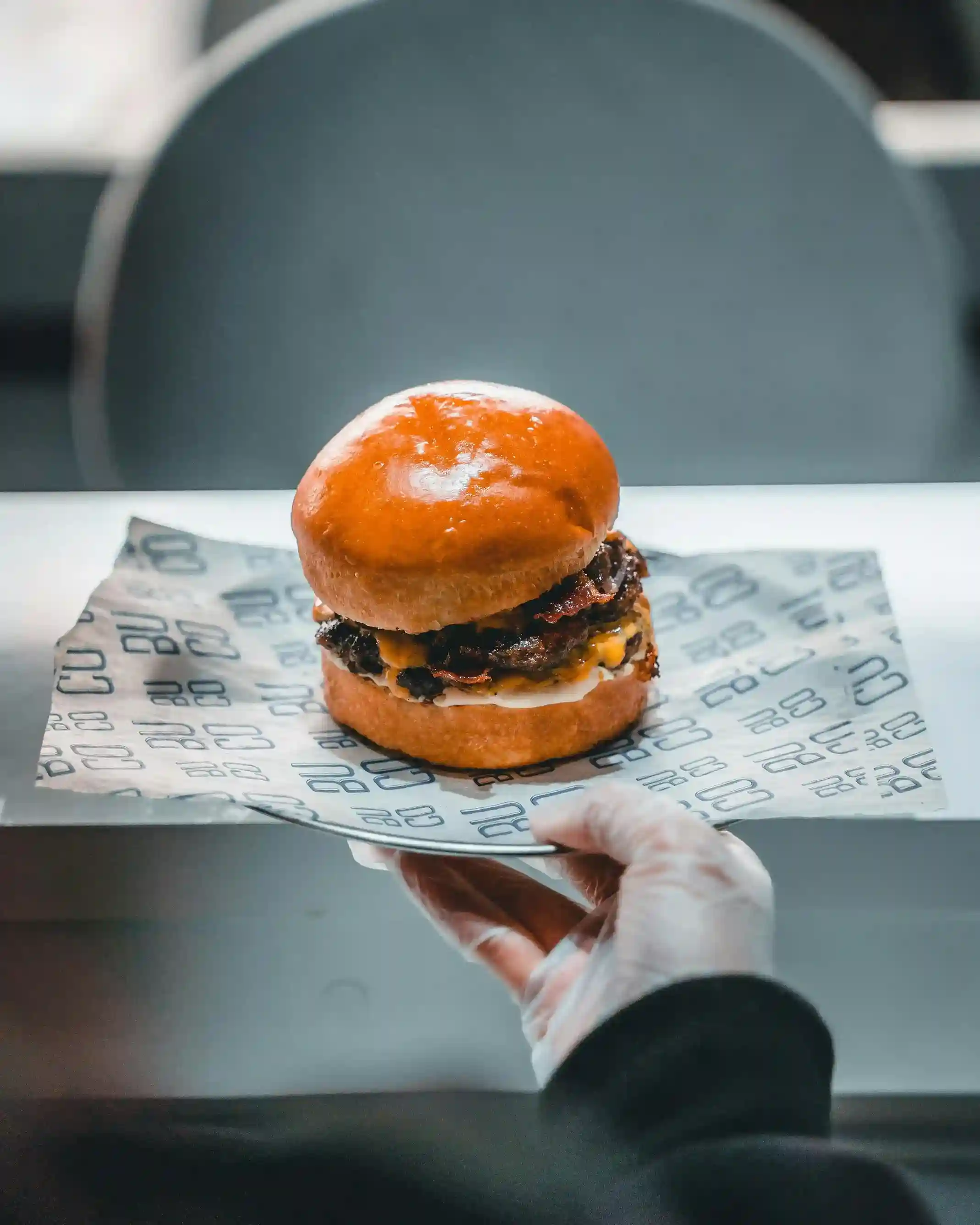 A gourmet burger served on custom-printed deli paper, featuring a glossy bun and melted cheese, held by a hand wearing gloves