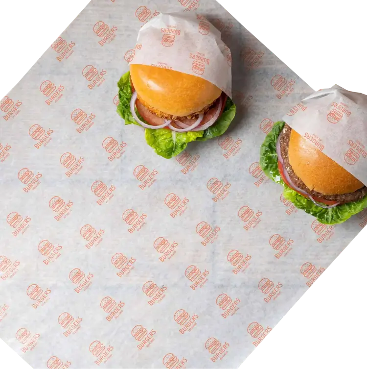 Two freshly made burgers wrapped in custom-printed deli paper with a burger logo design, placed on a matching deli paper sheet