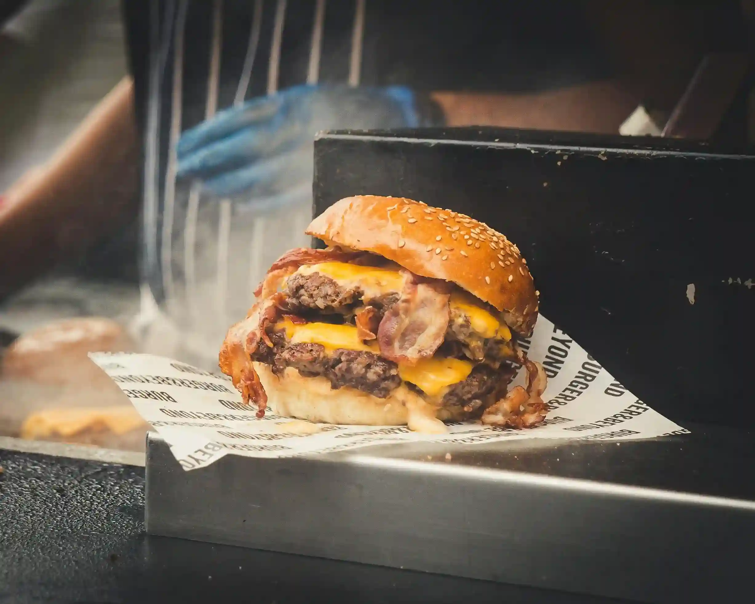 Juicy double bacon cheeseburger with sesame seed bun served on custom-printed food paper, prepared in a grilling station