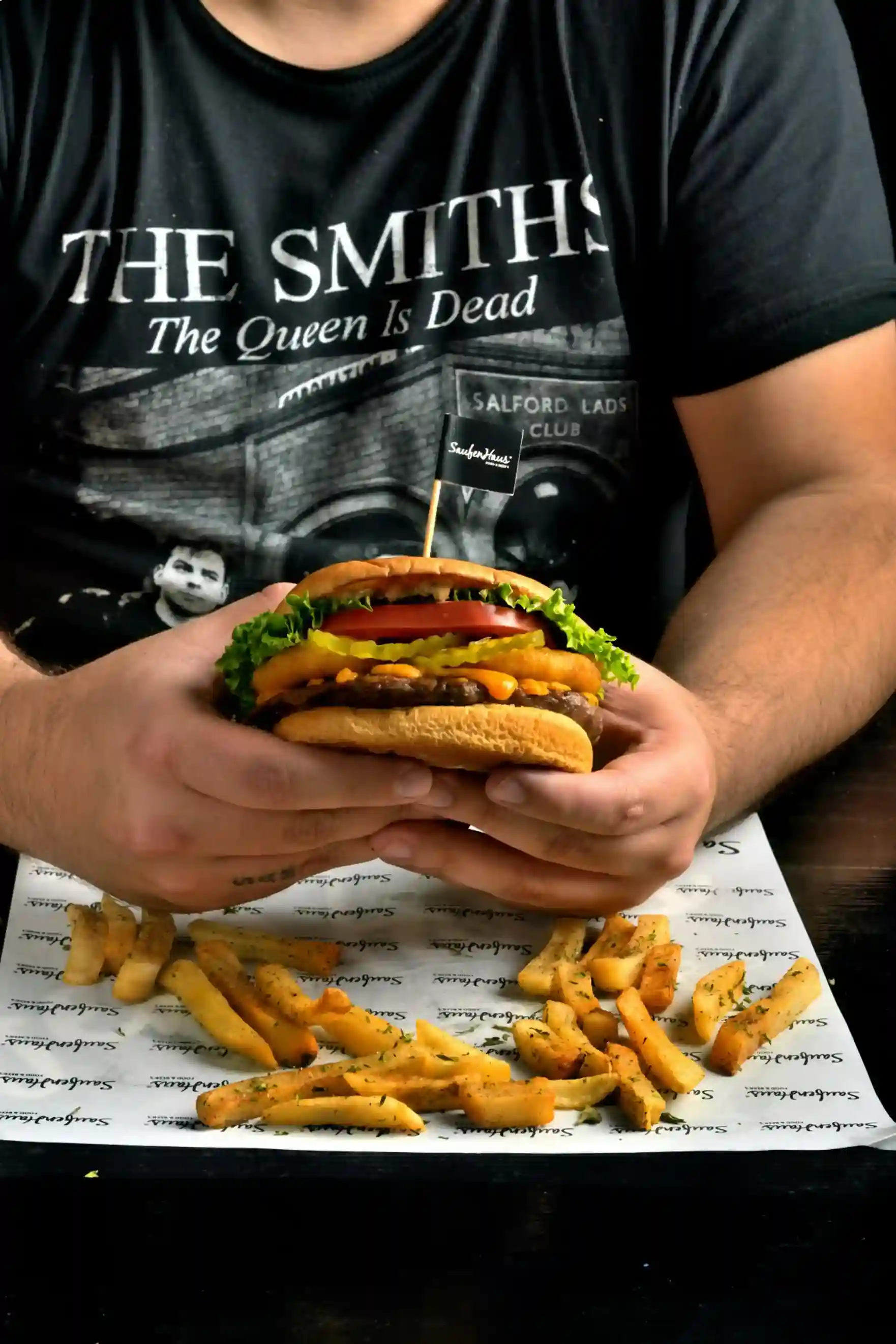 A person holding a burger with a flag toothpick, served with fries on custom-printed greaseproof paper featuring a branded design