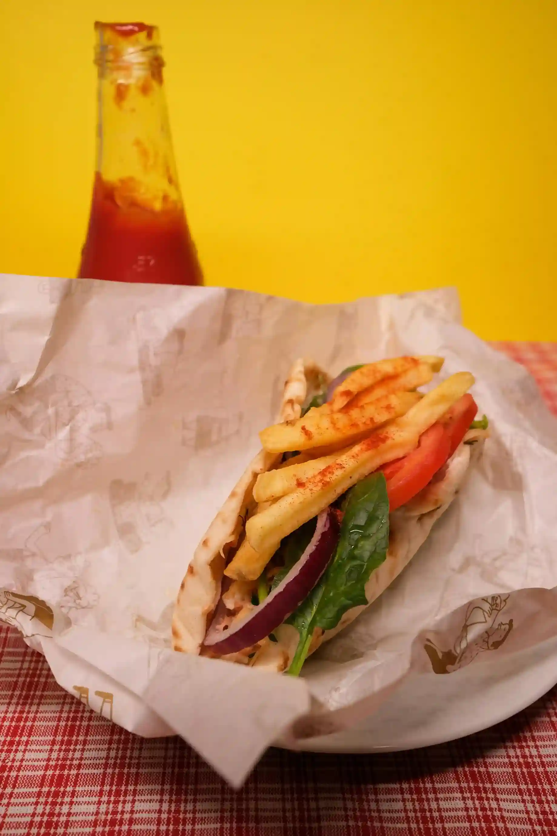 Gyro sandwich wrapped in custom hot paper, served on a plate with a bottle of ketchup in the background, placed on a red-checkered tablecloth for a vibrant presentation