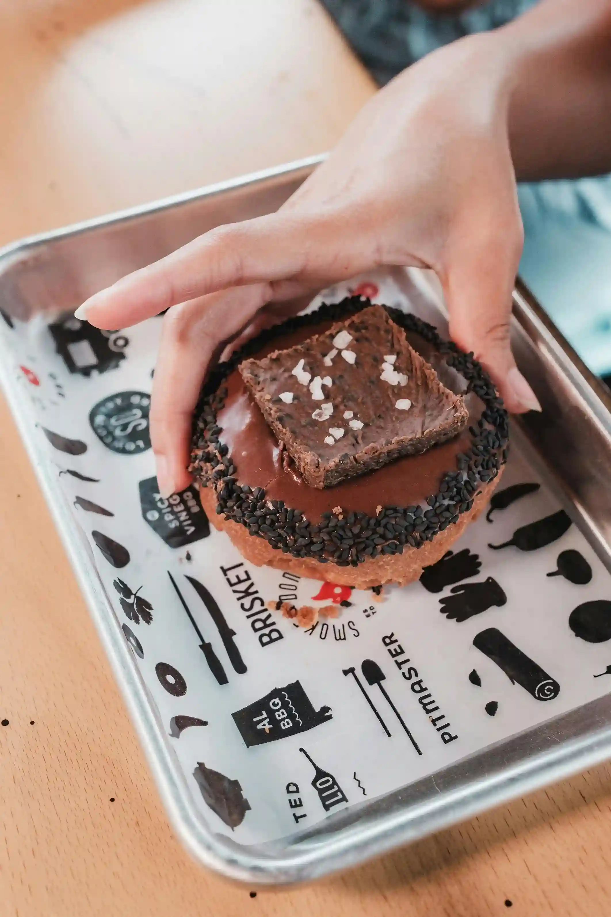 A hand holding a dessert topped with chocolate and sea salt, served on a tray lined with custom-printed parchment paper