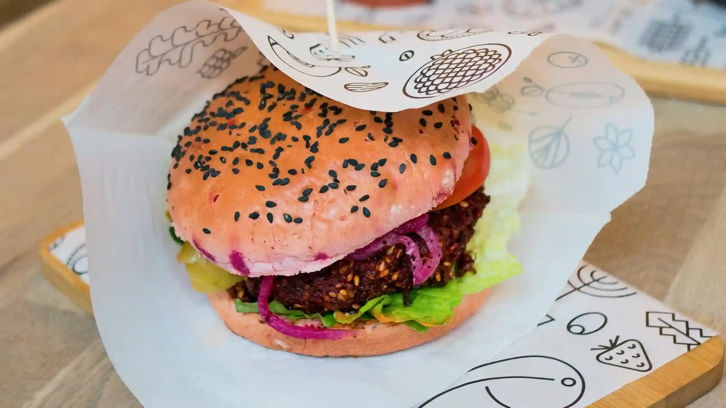 A gourmet burger with a sesame seed bun, wrapped in custom-printed parchment paper featuring minimalist food-themed illustrations