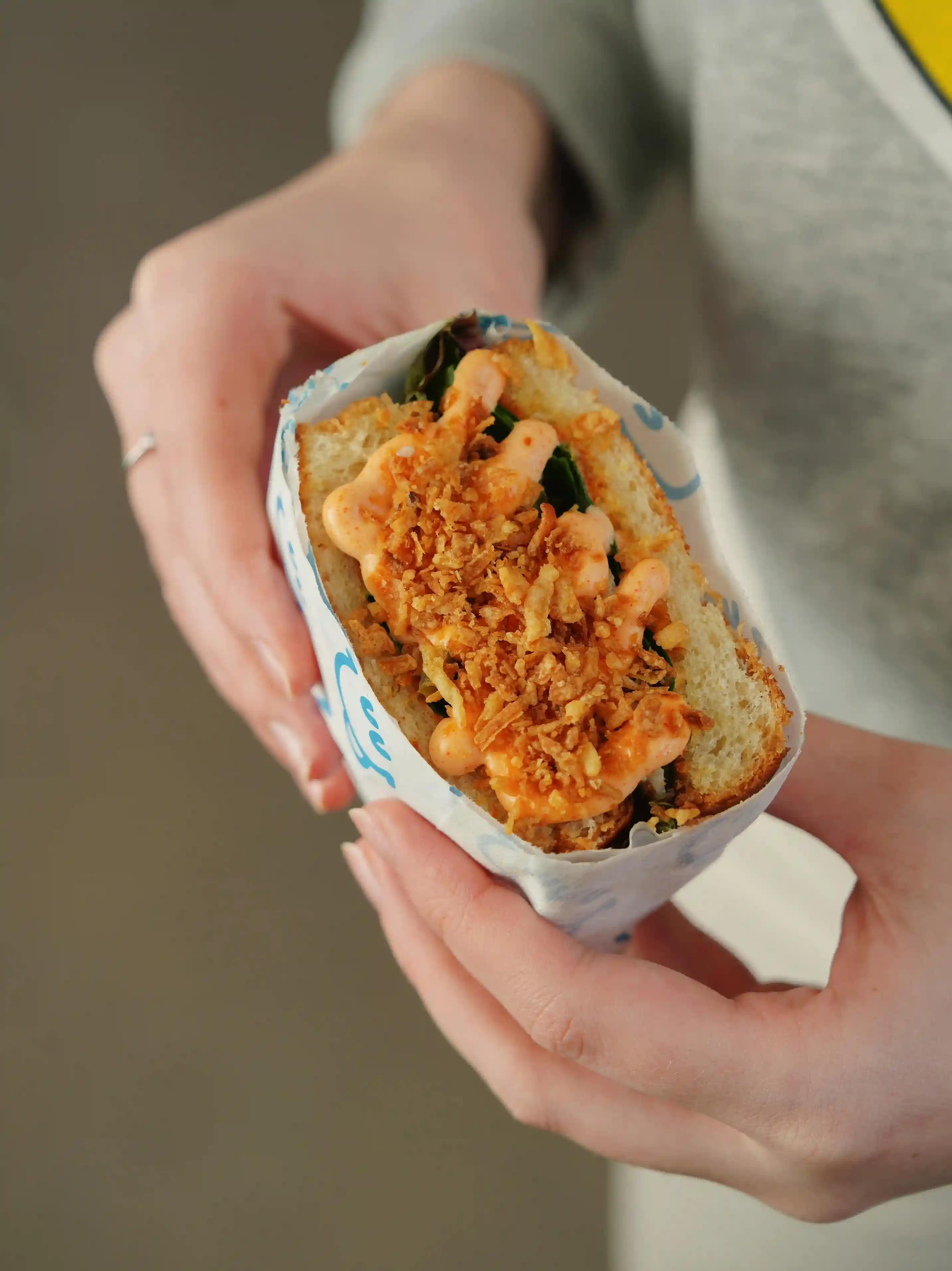 Close-up of a person holding a sandwich wrapped in custom-printed sandwich paper, showcasing a delicious filling topped with crispy fried onions and a creamy orange sauce