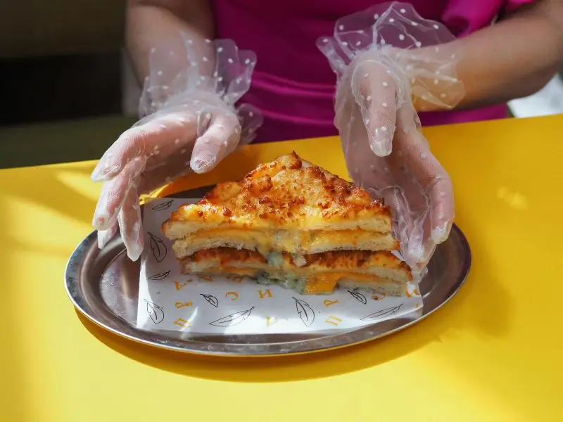 Gloved hands placing a sandwich onto custom printed wax paper in a tray. The wax paper is designed to prevent oil leaks and enhance food presentation