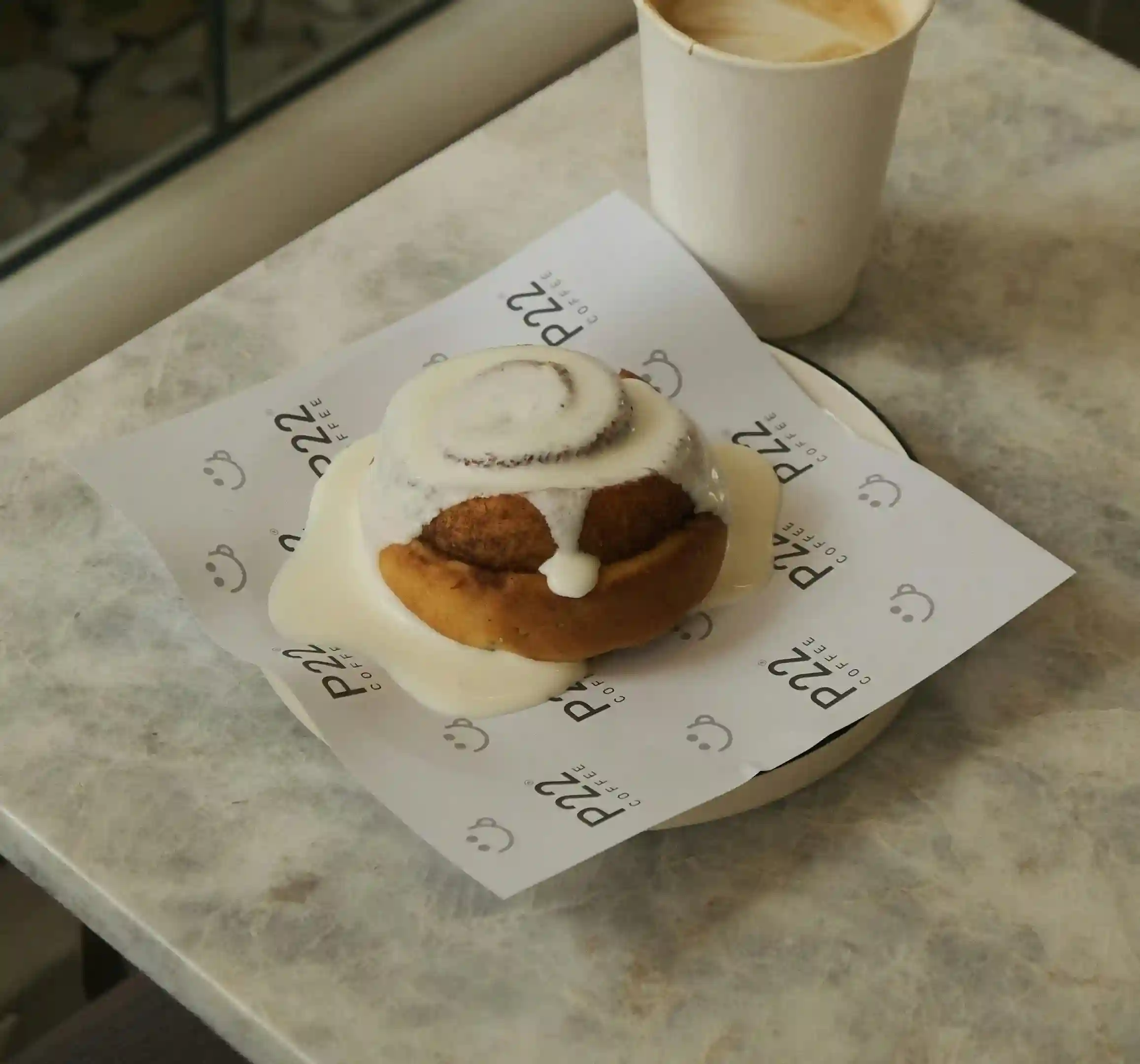 Warm cinnamon roll drizzled with icing, served on custom-printed parchment paper alongside a creamy latte in a paper cup